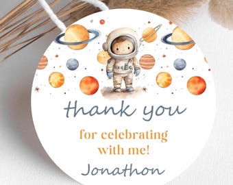 May include: Round thank you tag with a watercolor illustration of an astronaut in a white spacesuit standing on a white background with orange, yellow, and blue planets and stars. The text on the tag reads "Thank you for celebrating with me! Jonathon".