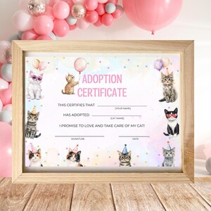 Puede incluir: Certificado de adopción enmarcado con el texto "ADOPTION CERTIFICATE" en rosa. El certificado presenta ilustraciones de acuarela de gatos con sombreros de fiesta y gafas de sol, con globos en el fondo. El marco es de madera clara.