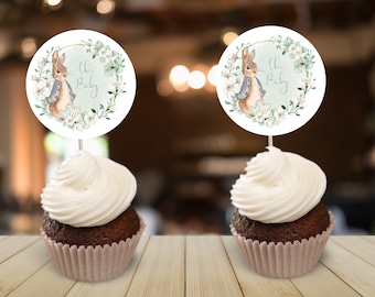 May include: Two cupcakes with white frosting and a brown base. Each cupcake has a round topper with a watercolor illustration of a bunny wearing a blue jacket and a wreath of green leaves and white flowers. The text "Oh Baby" is written on each topper.