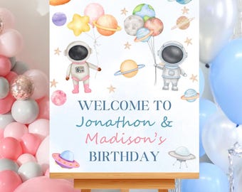 May include: A wooden easel displays a birthday sign with an outer space theme. The sign features illustrations of astronauts, planets, and stars, with the text "WELCOME TO Jonathon & Madison's BIRTHDAY". Balloons in pink, silver, and blue surround the sign.
