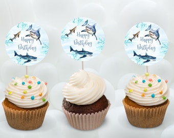 May include: Three cupcakes decorated with white frosting and colorful sprinkles. Each cupcake has a round blue and white paper topper with a shark illustration and the text "Happy Birthday".