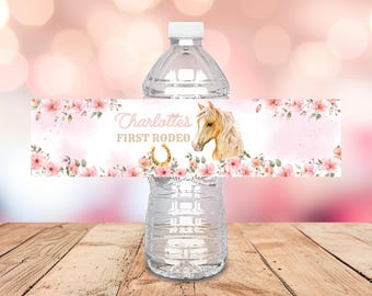 Op de afbeelding: Een doorzichtige waterfles met een aangepast etiket. Het etiket toont de tekst "Charlotte's FIRST RODEO" in roze script, een aquarel paardenkop, een gouden hoefijzer en roze bloemaccenten. De fles staat op een houten oppervlak.