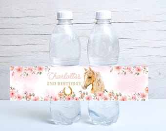 Op de afbeelding: Twee doorzichtige waterflessen met witte doppen en twee doorzichtige glazen. Een roze banner met bloemaccenten, een paardenillustratie en de tekst "Charlotte's 2nd Birthday" wikkelt zich om de flessen.