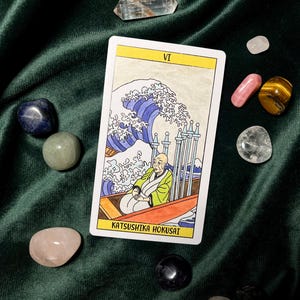 May include: A tarot card with a yellow border featuring an illustration of a man in a boat with a large wave. The card is surrounded by various polished gemstones, including clear quartz, rose quartz, and tiger's eye, on a dark green velvet surface.
