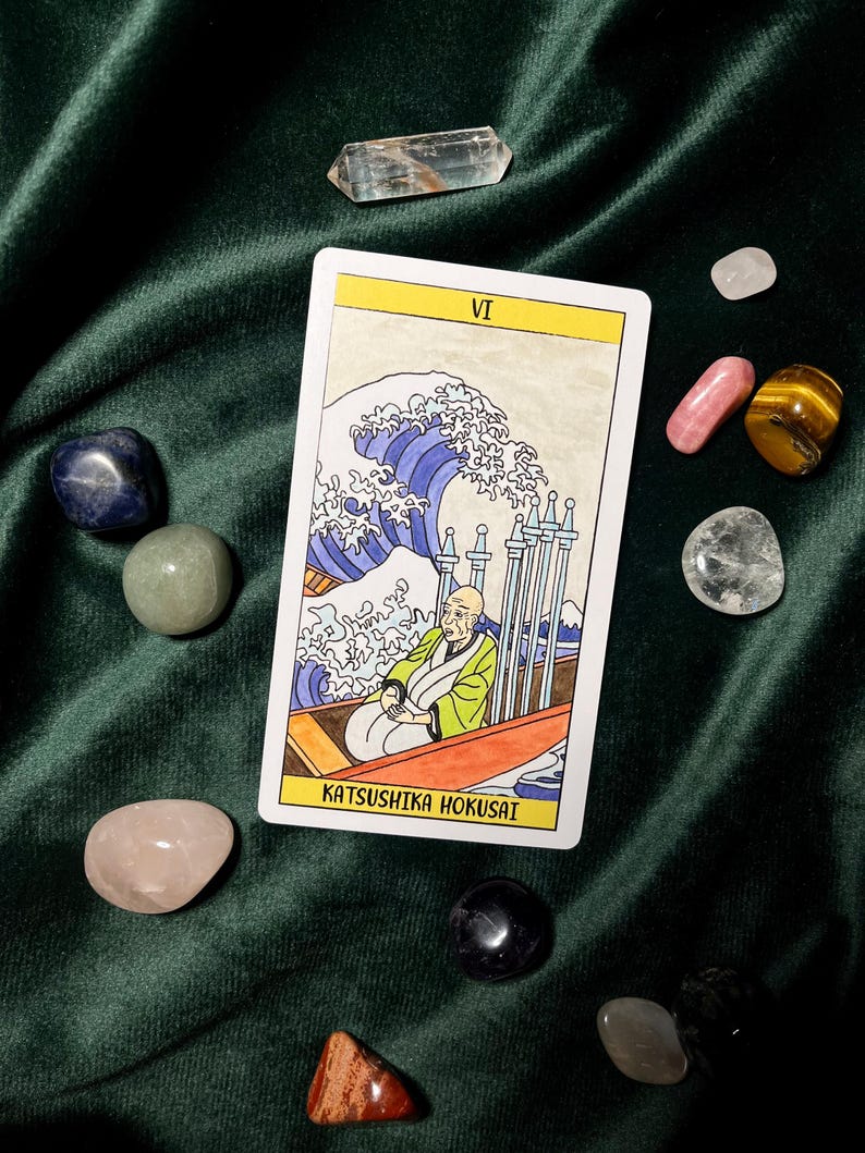 May include: A tarot card with a yellow border featuring an illustration of a man in a boat with a large wave. The card is surrounded by various polished gemstones, including clear quartz, rose quartz, and tiger's eye, on a dark green velvet surface.