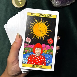May include: A tarot card from a deck featuring a colorful illustration of a woman in a red polka-dot dress, with a yellow sun and the words "THE MOON". The card is held with other cards.