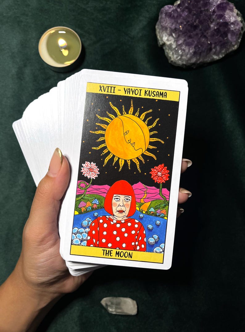 May include: A tarot card from a deck featuring a colorful illustration of a woman in a red polka-dot dress, with a yellow sun and the words "THE MOON". The card is held with other cards.
