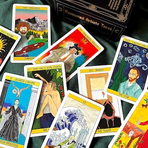 May include: A tarot card deck with the title "Renowned Artists Tarot" on the box. The cards feature illustrations of famous artists like Bob Ross, Jean-Michel Basquiat, and Vincent van Gogh. The cards are on a green surface.