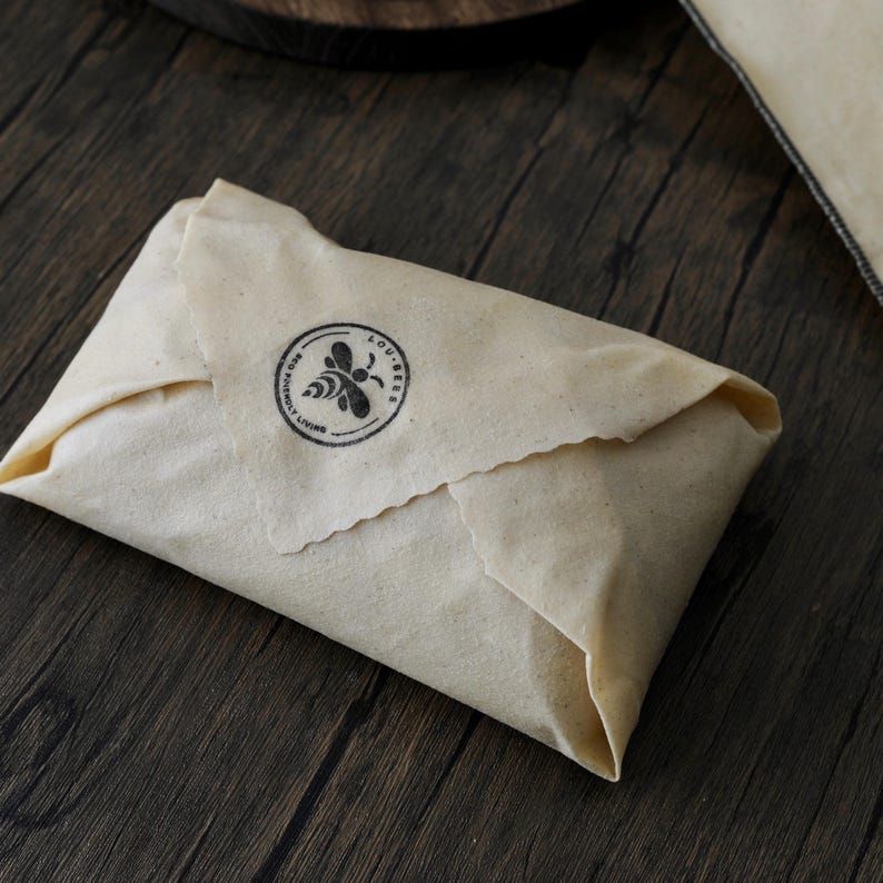 May include: A rectangular beeswax food wrap folded into a packet, made of off-white fabric with a visible texture. A circular black ink stamp on the top flap features a bee illustration and the text LOU-BERS ECO FRIENDLY LIVING.