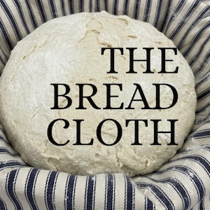 Könnte beinhalten: Ein rundes Brot, mit Mehl bestäubt, liegt in einem mit blau-weiß gestreiftem Stoff ausgekleideten Korb. Der Text "THE BREAD CLOTH" ist in großen, schwarzen Buchstaben auf das Brot gedruckt.
