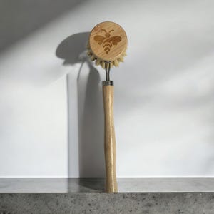 Puede incluir: Un cepillo de madera para lavar platos con una cabeza redonda con un diseño de abeja. El cepillo tiene cerdas naturales y un mango largo de madera clara. El diseño de la abeja está grabado en la madera. El cepillo está diseñado para lavar platos.