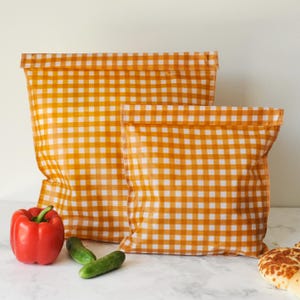 May include: Two rectangular food storage bags featuring a vibrant orange and white gingham check pattern. The bags are constructed with a flat, folded top edge and a flexible, lightweight material, displayed alongside a red bell pepper and cucumbers.