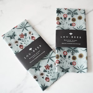 May include: Two light blue beeswax wraps with floral patterns, featuring red, white, and yellow flowers, and green leaves. Each wrap has a black band with the text "LOU-BEES Beeswax wraps Natural, reusable food storage."