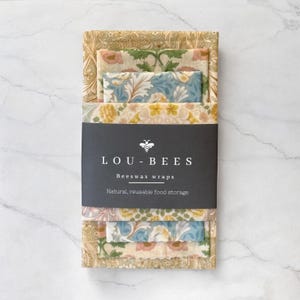 May include: A set of patterned beeswax wraps in various sizes. The wraps feature floral designs in shades of pink, blue, yellow, and green. The wraps are held together with a black label that reads "LOU-BEES Beeswax wraps Natural, reusable food storage."
