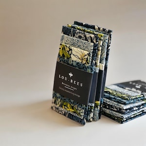 Może przedstawiać: Stos wzorzystych woskowanych opakowań z czarną etykietą z napisem "LOU-BEES" i "Beeswax wraps". Opakowania mają kwiatowy wzór w odcieniach niebieskiego, zielonego i białego. Opakowania służą do naturalnego, wielokrotnego użytku przechowywania żywności.
