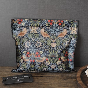 Pode incluir: Um saco quadrado de cera de abelha para alimentos com fundo azul escuro e um padrão repetitivo de pássaros castanhos, folhagem verde e morangos vermelhos. Uma etiqueta retangular preta com o texto THE BEESWAX BAG está em primeiro plano.