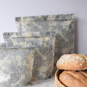 Puede incluir: Tres bolsas reutilizables para guardar alimentos con un estampado floral en azul y crema. Las bolsas están hechas de un material flexible y están dobladas en la parte superior. Una cesta de pan está en el fondo.