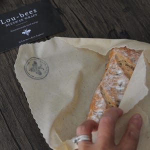 May include: A beige beeswax food wrap with a circular bee logo stamp, partially covering a loaf of bread. A black rectangular card is visible with the white text Lou-bees BEESWAX WRAPS Natural reusable food storage by Lou-bees Kent, England.