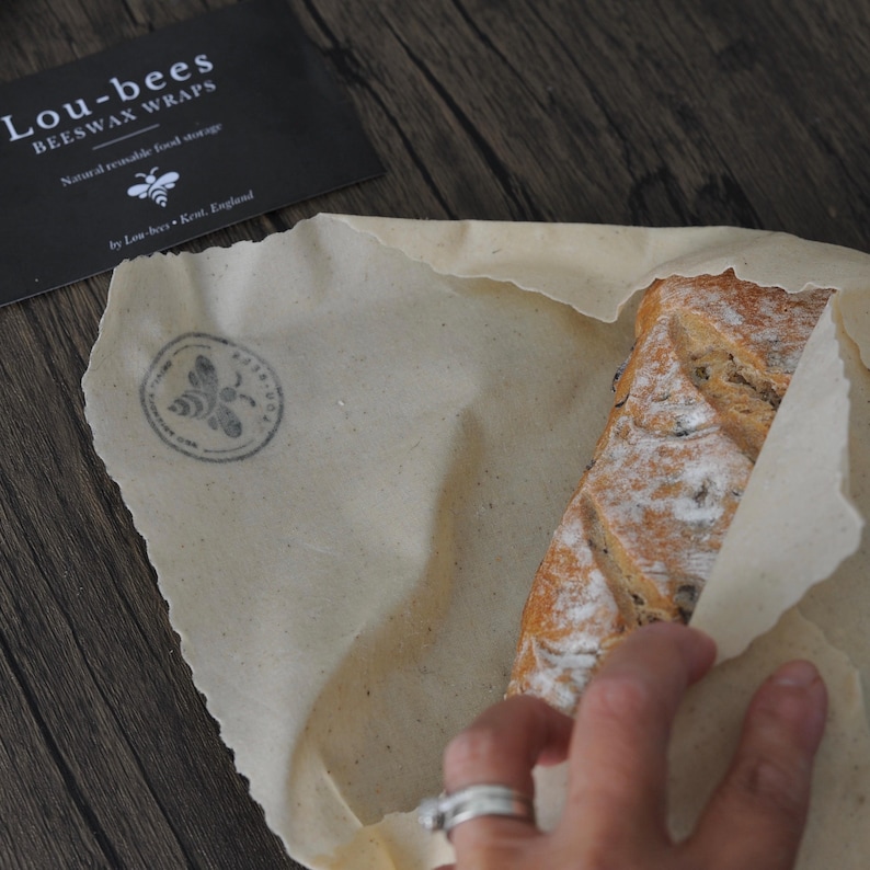 May include: A beige beeswax food wrap with a circular bee logo stamp, partially covering a loaf of bread. A black rectangular card is visible with the white text Lou-bees BEESWAX WRAPS Natural reusable food storage by Lou-bees Kent, England.