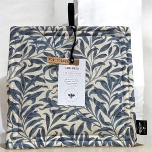 May include: A square pot holder featuring a blue and white leaf design. It has a black hanging loop and a tag that reads "POT HOLDER" and "LOU-BEES". The tag also includes information about the materials used.