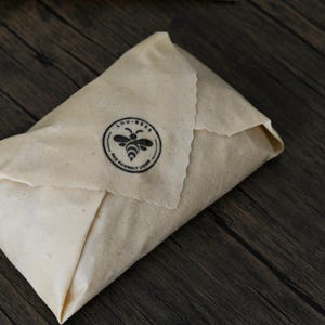May include: A rectangular piece of beige beeswax food wrap folded into a packet. The wrap features a black circular stamp with a bee illustration in the center and the text LOU-BEES ECO FRIENDLY LIVING printed around the bee icon.