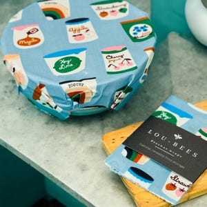 Puede incluir: Un juego de envoltorios de alimentos de cera de abeja azul y blanco con un patrón colorido de envases de yogur. Los envoltorios están hechos por LOU-BEES y son un almacenamiento de alimentos natural y reutilizable.