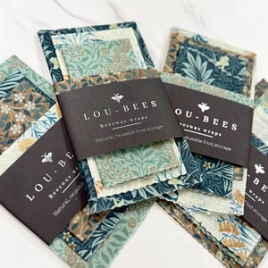 May include: Several sets of patterned beeswax wraps are displayed, each wrapped with a black paper band. The wraps feature floral designs in shades of blue, green, and brown. The text "LOU-BEES Beeswax wraps" is printed on the bands.
