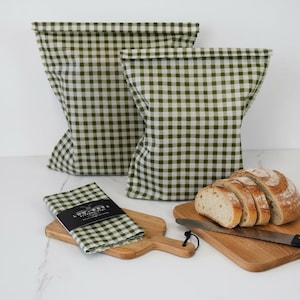 May include: Two olive green and white gingham fabric bags with folded tops, a folded matching gingham cloth with a black label reading LOU BARK, and a sliced loaf of bread on a wooden cutting board with a serrated knife.