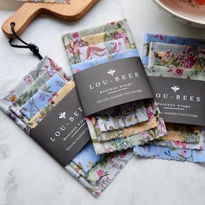 May include: Several stacks of floral patterned beeswax wraps are displayed. Each stack is wrapped with a black label that reads "LOU-BEES Beeswax wraps Natural, reusable food storage". The wraps feature various pastel colors and floral designs.