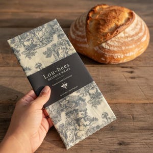 May include: A rectangular beeswax food wrap featuring a cream background with a dark gray toile de jouy pattern. A black paper band is wrapped around the center with the white text Lou-bees BEESWAX WRAPS and a small bee icon printed on it.