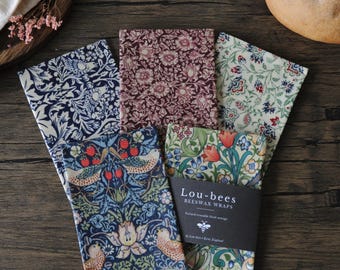 May include: A set of five rectangular beeswax wraps featuring various floral and bird patterns in shades of blue, red, green, and cream. A black paper band labeled Lou-bees BEESWAX WRAPS is wrapped around the center of the collection.