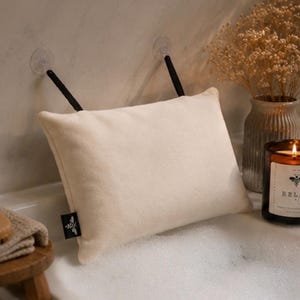 Puede incluir: Una almohada de baño rectangular de color crema con dos correas negras y ventosas transparentes para su fijación. En el lateral de la almohada hay una pequeña etiqueta de tela negra con el icono de una abeja en color blanco.