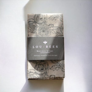 Puede incluir: Un paquete de tres envoltorios de alimentos de cera de abeja con un patrón floral. Los envoltorios están empaquetados con una etiqueta negra que dice "LOU-BEES Beeswax wraps Natural, reusable food storage".