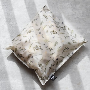 Pode incluir: Um envoltório de comida reutilizável branco com um padrão floral de folhas verdes e bagas roxas. O envoltório tem um logotipo de abelha preto no canto inferior direito.