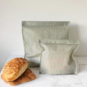 Puede incluir: Dos bolsas de pan de tela a rayas verdes y blancas de diferentes tamaños están junto a dos hogazas de pan. El pan tiene una corteza dorada. Las bolsas están dobladas en la parte superior.