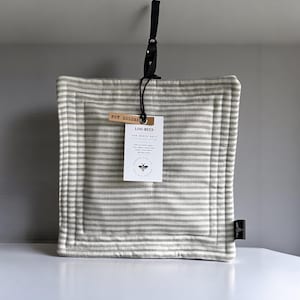 May include: A square, gray and white striped pot holder with a black leather loop for hanging. The pot holder has a tag that reads "POT HOLDER" and "LOU-BEES". The pot holder is made of cotton.