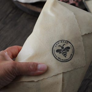 May include: A beige, flexible food wrap with a textured surface and pinked edges. A circular black stamp is printed on the material, featuring a stylized bee icon and the text LOU-BEES ECO FRIENDLY LIVING arranged in a circular border.