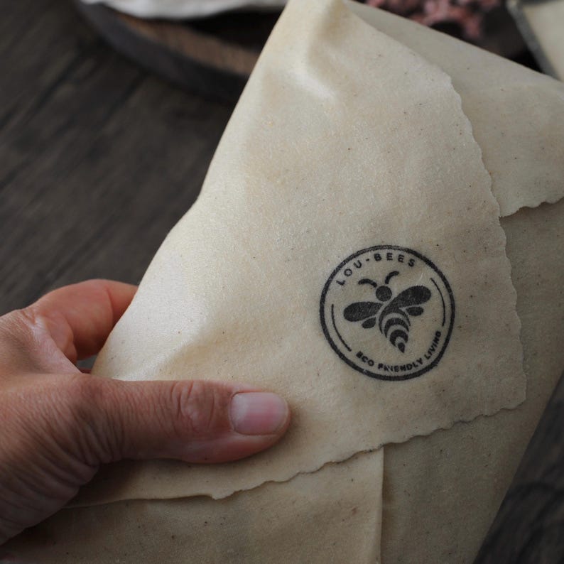 May include: A beige, flexible food wrap with a textured surface and pinked edges. A circular black stamp is printed on the material, featuring a stylized bee icon and the text LOU-BEES ECO FRIENDLY LIVING arranged in a circular border.