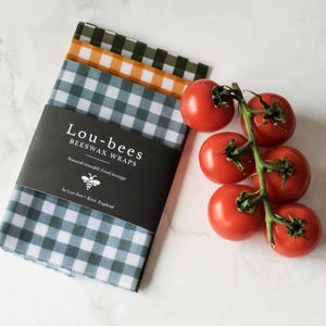 May include: A stack of three folded beeswax wraps featuring a blue, orange, and dark green gingham pattern. A black paper band wraps around the center with the white text Lou-bees BEESWAX WRAPS, a bee icon, and the text by Lou-bees Kent, England.