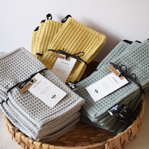 Puede incluir: Paños de cocina de tejido de gofre en gris, amarillo y verde, atados con hilo negro y etiquetas, se exhiben en una cesta de mimbre. Las etiquetas dicen "Dishcloth" y "Lou-Bees". La imagen presenta una disposición ordenada y organizada.