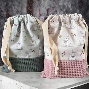 Puede incluir: Dos bolsas de cordón con un patrón de conejo. La bolsa de la izquierda es verde y la bolsa de la derecha es rosa.