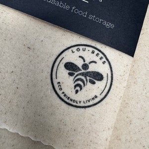 May include: A beige fabric beeswax wrap with a scalloped edge, featuring a black circular logo with a bee illustration and the text LOU-BEES ECO FRIENDLY LIVING. A black paper label at the top reads LOU-BEES Beeswax wraps Natural, reusable food storage.