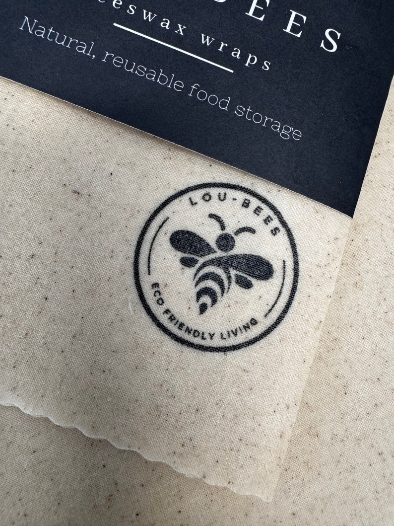 May include: A beige fabric beeswax wrap with a scalloped edge, featuring a black circular logo with a bee illustration and the text LOU-BEES ECO FRIENDLY LIVING. A black paper label at the top reads LOU-BEES Beeswax wraps Natural, reusable food storage.