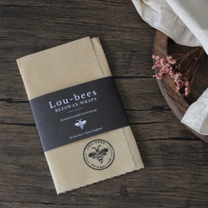 May include: A rectangular, light tan beeswax wrap with scalloped edges, featuring a black paper band printed with the white text Lou-bees BEESWAX WRAPS, Natural reusable food storage, by Lou-bees Kent, England, and a bee logo icon.