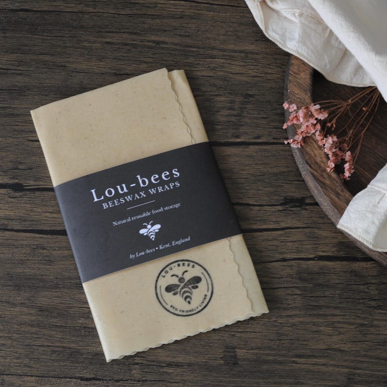 May include: A rectangular, light tan beeswax wrap with scalloped edges, featuring a black paper band printed with the white text Lou-bees BEESWAX WRAPS, Natural reusable food storage, by Lou-bees Kent, England, and a bee logo icon.