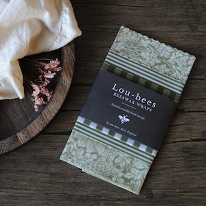 May include: A stack of folded beeswax wraps featuring sage green floral and striped patterns, secured by a black paper band labeled Lou-bees BEESWAX WRAPS Natural reusable food storage by Lou-bees Kent, England with a small bee icon.