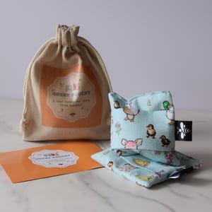 Peut inclure: Un "Ouchy Pouchy" bleu clair avec un motif de canards et de cochons, un sac à cordon et une carte orange. La pochette est conçue pour offrir une douce étreinte pour les petits tracas de la vie. Le sac porte l'inscription "Kids Ouchy Pouchy".