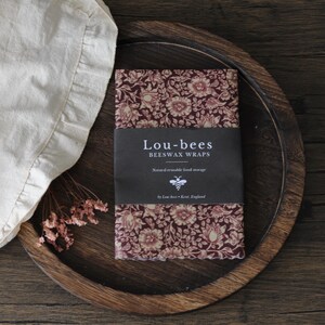 May include: A rectangular beeswax wrap featuring a dark red and cream floral pattern. A black paper band wraps around the center with the white text Lou-bees BEESWAX WRAPS, Natural reusable food storage, and a small bee icon by Lou-bees Kent, England.