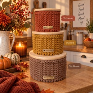 Op de afbeelding: Een stapel van drie ronde, gehaakte opbergmanden met lichtgekleurde houten deksels. De bovenste mand is terracotta, de middelste is mosterdgeel en de onderste is nootbruin. Elke mand heeft een klein rechthoekig metalen label met HM Design.