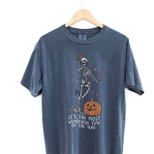 Könnte beinhalten: Zwei T-Shirts mit einem Skelett-Motiv und dem Text "It's the most wonderful time of the year". Das Shirt links ist orange und das Shirt rechts ist blau.